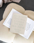 The beige "101 Quotes That Will Change the Way You Think" by Faire rests atop an open book on a light cushioned seat, providing daily inspiration with selected quotes.