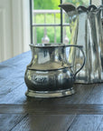 The Vintage 1815 Large Pitcher by Faire, crafted in Italy, sits on a dark wooden table near a window, with two other metal pitchers behind it as bright natural light streams in.