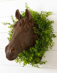 The Faire Horse Head Wall Mount is displayed on a white shiplap wall, accented by a green leafy wreath that forms a decorative frame around the rustic brown sculpture.