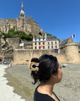 A woman with dark hair in a Faire Fashion Hair Clip stands on sandy ground, looking at Mont Saint-Michel’s historic stone building with flags, under a blue sky.