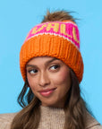 A woman with long brown hair wears a tan sweater and a Faire Knit Ski Hat—an orange luxury knit beanie with "CHILL" in pink and white—against a solid light blue background, smiling softly and looking slightly to the side.