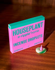 A pink and green box labeled "Houseplant by Ripple/ Incense Droplets" by Faire sits on a wooden surface next to a modern incense holder with a burning pink cone, releasing refreshing home fragrance as smoke swirls upward.