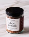 A glass jar with a black lid, labeled "Faire Gaby's Everyday," sits on a white marble surface, filled with a flavorful blend of herbs and spices like garlic and smoky paprika.