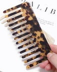 A hand holds the Faire Hair Comb Assorted, featuring a wide-toothed, anti-static design and tortoiseshell pattern, over an open magazine with visible black text in the background.