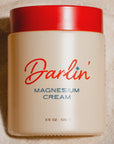 A beige jar of Magnesium Cream with a red lid, labeled "Magnesium Cream 3.5 oz / 100 g," is photographed from above as it rests on sand.