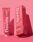 A pink tube and box of Faire Davids Toothpaste in naturally sweet strawberry watermelon flavor stand upright on a pink background. The fluoride-free toothpaste comes in a recyclable metal tube with bold white text on the packaging.