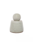 The Faire Alabaster White Stash Jar is a small, handmade stoneware container with a rounded lid. It features a matte light gray glaze with brown speckles, highlighting its minimalist design, and sits on a white background.
