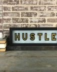 A Faire Lighted Sign with "HUSTLE" in bold black and gold letters rests on a wooden table beside stacked books and a white teacup, all set against a brick wall.