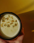 Faire Gold Dust Woman Candle with a creamy gold-flecked drink, styled on a vibrant yellow surface for Bungalow ambiance.