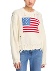 Wearing Denimist’s OVERSIZED CROPPED AMERICAN FLAG SWEATER—an off-white crewneck featuring a large American flag, frayed edges, and distressed holes—paired with blue jeans for a worn-in look.