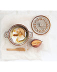 A round Brie cheese melts in a Creative Co-op Brie Baker with "BRIE BAKER" on the lid. Sliced pear tops the cheese, and a halved pear and bamboo spreader rest nearby on white parchment paper.