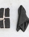 Two charcoal stonewashed linen napkins from the Creative Co-op S/4 set are displayed on a light textured surface; one is neatly folded and tied with cotton tape, the other is unfolded and draped alongside.