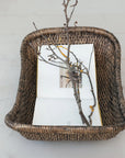 The Creative Co-op Woven Wicker Vintage basket cradles an open book topped with a dried branch, displayed on a light floor for an antique touch in a tranquil setting.