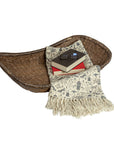 The Creative Co-op Woven Wicker Vintage basket, with an antique finish, holds a cream floral throw, two stacked books, and eyeglasses, all displayed against a white background.