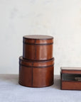 Two round DG0045 Leather Boxes by Creative Co-op, in cognac MDF & leather, are stacked beside a rectangular leather and glass tray with rope on a light surface against a neutral background.