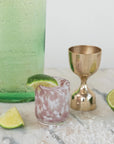 A marbled shot glass with a drink and lime wedge rests on marble next to the Creative Co-op Brass Double Sided Cocktail Jigger, a green bottle, lime slices, and coarse salt.