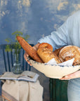 A person in a light blue shirt holds the Creative Co-op Round 15" Terra-cotta Bowl filled with assorted artisan breads. Behind them, a chair with a folded cloth, open book, and greenery vase rests against a textured blue wall.