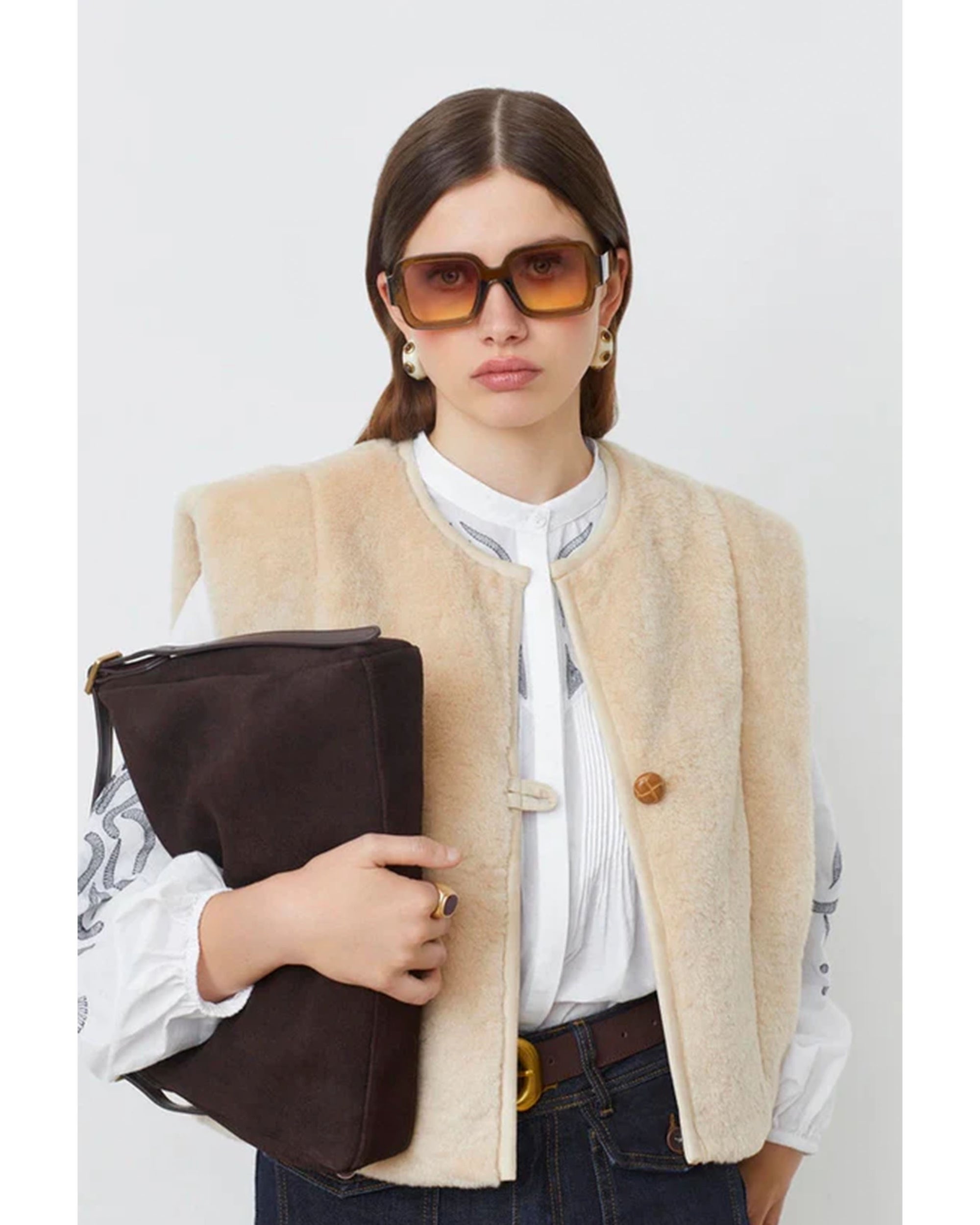 A woman with straight brown hair wears SUNCOO's Diego Vest over a white blouse, paired with blue jeans, gold earrings, and large square sunglasses. She holds a large brown clutch against a plain white background.