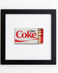 A framed piece of art displaying a matchbox designed to look like a Diet Coke can. The Art Square Blk Frame, by Match South, with its gray and red color scheme, contains several red-tipped matches. Enclosed in clear acrylic, the black frame contrasts beautifully against the white background.