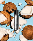 A bottle of Faire Heretic 15ML Perfume rests on a blue surface, surrounded by cracked open coconuts and pieces of coconut flesh.