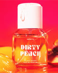 A clear bottle of Heretic 50ML Perfume by Faire, labeled "Dirty Peach," rests against a vibrant red and orange background with peach slices and syrup, suggesting a sensual fragrance featuring sandalwood vanilla notes.