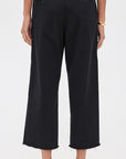 A person wears Denimist's CROPPED DOUBLE PLEAT WIDE LEG CHINO in black 100% cotton with raw hems, paired with a white top, black shoes, and gold jewelry, shown from the back against a plain white background.