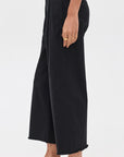 A person wearing Denimist CROPPED DOUBLE PLEAT WIDE LEG CHINO in black, paired with a white top, black loafers, and gold rings, stands against a plain light background.