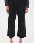 A person stands against a plain white background wearing a chunky gray knit sweater, Denimist CROPPED DOUBLE PLEAT WIDE LEG CHINO pants in black, black loafers, and gold bracelets.