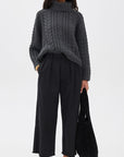 A woman stands against a plain white background wearing a chunky dark gray cable knit turtleneck, Denimist CROPPED DOUBLE PLEAT WIDE LEG CHINO pants in black, black loafers, sunglasses, and holding a black tote bag.