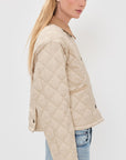 A woman with long blonde hair models the Denimist FAULKNER QUILTED JACKET, a beige lightweight jacket with snap closures, and light blue jeans, shown in a side pose against a plain white background.