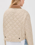 A woman with long blonde hair is seen from behind wearing the Denimist FAULKNER QUILTED JACKET—a beige quilted jacket with snap closure and brown collar—over a white shirt and light blue jeans, set against a plain white background.