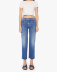 A woman stands facing forward against a white background, wearing Mother’s The Mid Rise Rambler Zip Ankle Fray jeans, a short-sleeved white crop top, and dark blue slip-on sandals.