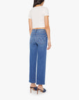 A woman wears Mother’s The Mid Rise Rambler Zip Ankle Fray jeans, a white short-sleeve top, and blue heeled shoes, standing with her back to the camera against a plain white background.