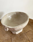The Tiny Trophy Bowl by Faire is a handmade, off-white glazed stoneware piece featuring an uneven rim and small circular handle, displayed on wood against a white wall—a unique sugar dish.