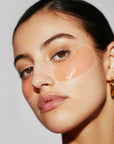 A woman with sleek dark hair and gold hoop earrings wears the Faire Hydrogel Eye Mask to hydrate and reduce wrinkles, looking confidently at the camera against a plain light background.