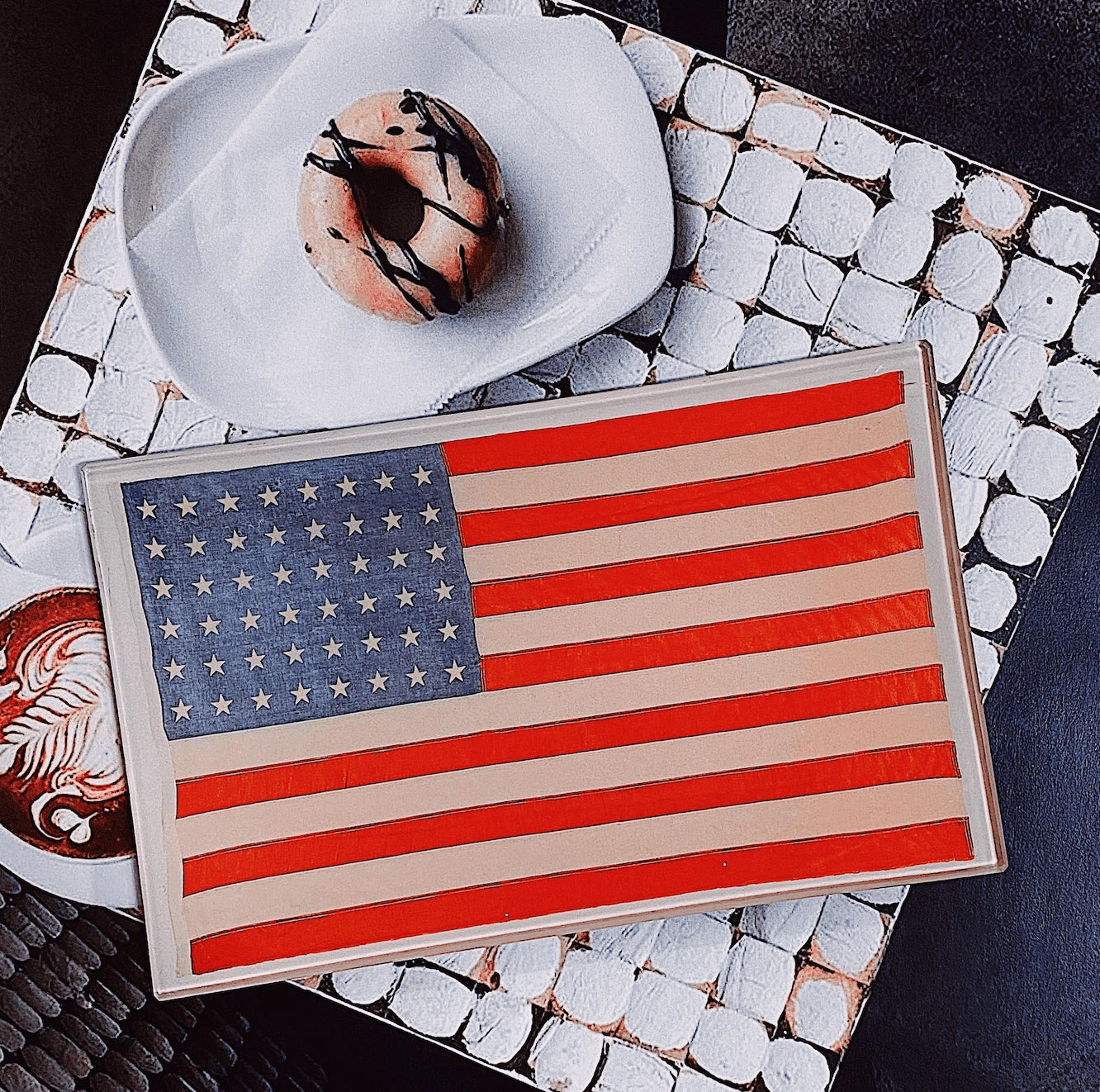 An American Flag cake rests on the Faire American Flag Glass Tray, beside a donut with chocolate drizzle and a latte. The geometric black and white table highlights the patriotic decor.