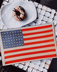 An American Flag cake rests on the Faire American Flag Glass Tray, beside a donut with chocolate drizzle and a latte. The geometric black and white table highlights the patriotic decor.