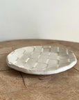The Faire Ghost Checker Trinket Dish, a small, round glazed stoneware dish with a woven checkerboard pattern, rests on a wooden surface against a plain, light-colored background.