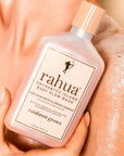 A close-up shows a person holding a pink bottle of Faire Rahua Enchanted Island Body Glow Wash against their soapy shoulder, with the moisturizing body wash’s label clearly visible.