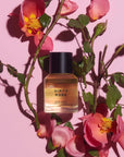 A bottle of Faire Heretic 50ML Perfume, labeled “Dirty Rose,” is displayed amid pink rose flowers and green leaves on a pink background, evoking a sensual fragrance.