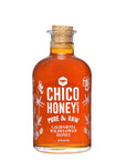 A clear glass bottle of Olivarez Chico Honey by Faire with a cork stopper, featuring white floral designs and text on the front label.