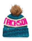The Faire Knit Ski Hat is a luxury blue, teal, and white merino wool beanie featuring a large brown faux fur pom-pom and bold pink "JACKSON" lettering across the front.