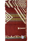 The Achuar Blanket by Faire is a folded Ecuadorian blanket with red, cream, and green geometric patterns—zigzags, diamonds, and arrows—and a small white label in one corner.