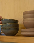 A hand reaches for the top Indaba Rowe Bowl Moss in a stack of three green reactive-glaze stoneware bowls on a wooden shelf, beside another stack of four bowls and plates, set against a light wood background.
