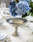 The Vintage 1795 Pedestal Bowl by Faire rests on a cream tablecloth, surrounded by white dishes, silverware, glassware, and a vase filled with blue hydrangeas and green leaves.