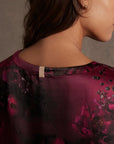 A woman with long brown hair faces away, revealing her bare neck and shoulder in a deep burgundy Lunya Washable Silk Tee by Faire, featuring a floral pattern and small beige neckline tag against a softly lit neutral background.