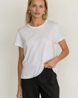 A woman with light hair wears Donni’s The Jersey Tee in plain white Supima cotton and dark pants, standing against a neutral background with a relaxed expression and one hand partly in her pocket.