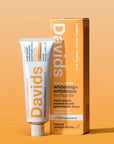 A tube and box of Faire Davids Toothpaste stand upright against an orange background. The recyclable metal tube highlights whitening, antiplaque, and fluoride-free benefits for kids and adults.