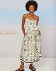 A woman smiles in the Me369 Elizabeth Midi Skirt, a cream, pleated piece with black ocean doodles and "TAKE ME TO THE OCEAN" lettering, creating a breezy summer vibe against a pale beach backdrop.