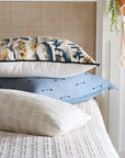 A stack of three pillows on a bed: the top is the Sprouse Watercolor pillow by Eastern Accents, followed by a blue cotton pillow with stitched dots and a cream geometric knit pillow. A hanging plant is visible on the right.